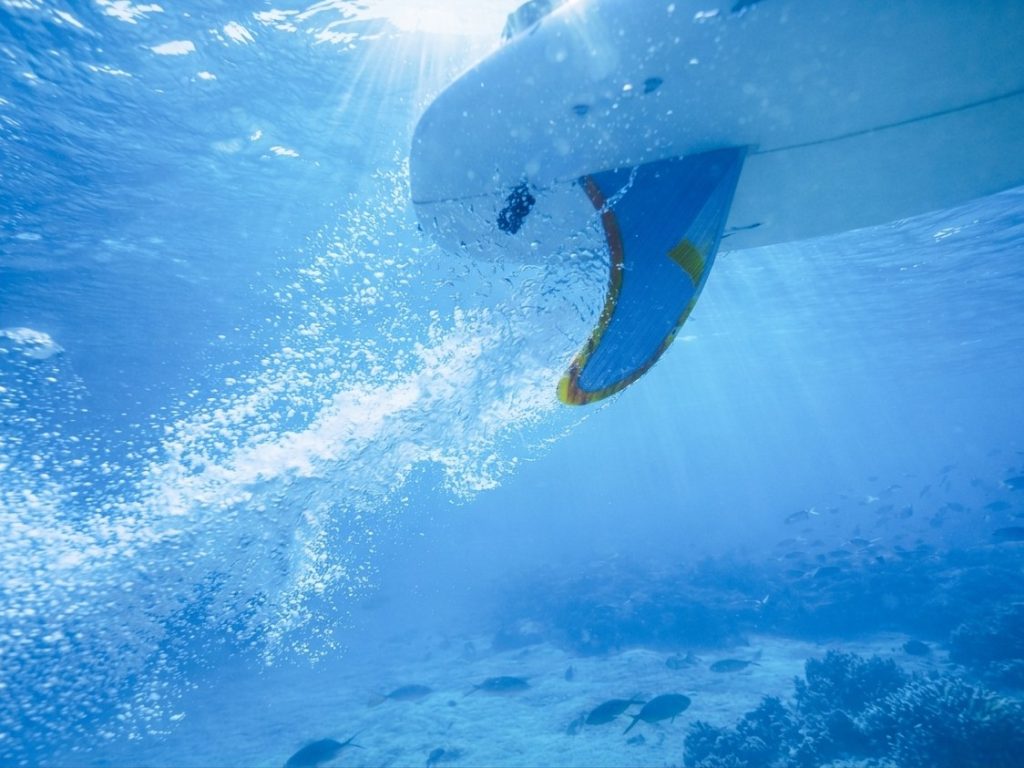 Sunlit underwater view of a surfboard and fin gliding through clear blue water above a reef